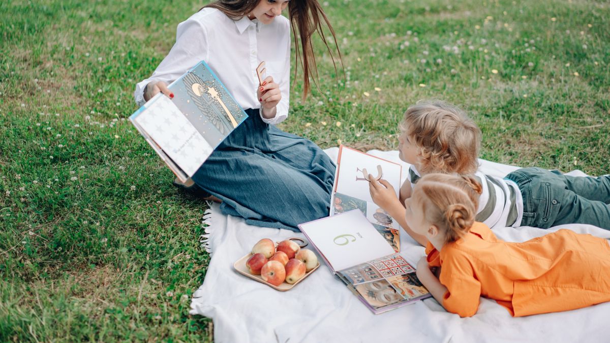 An adult reading textbooks outdoors with two children on the grass
