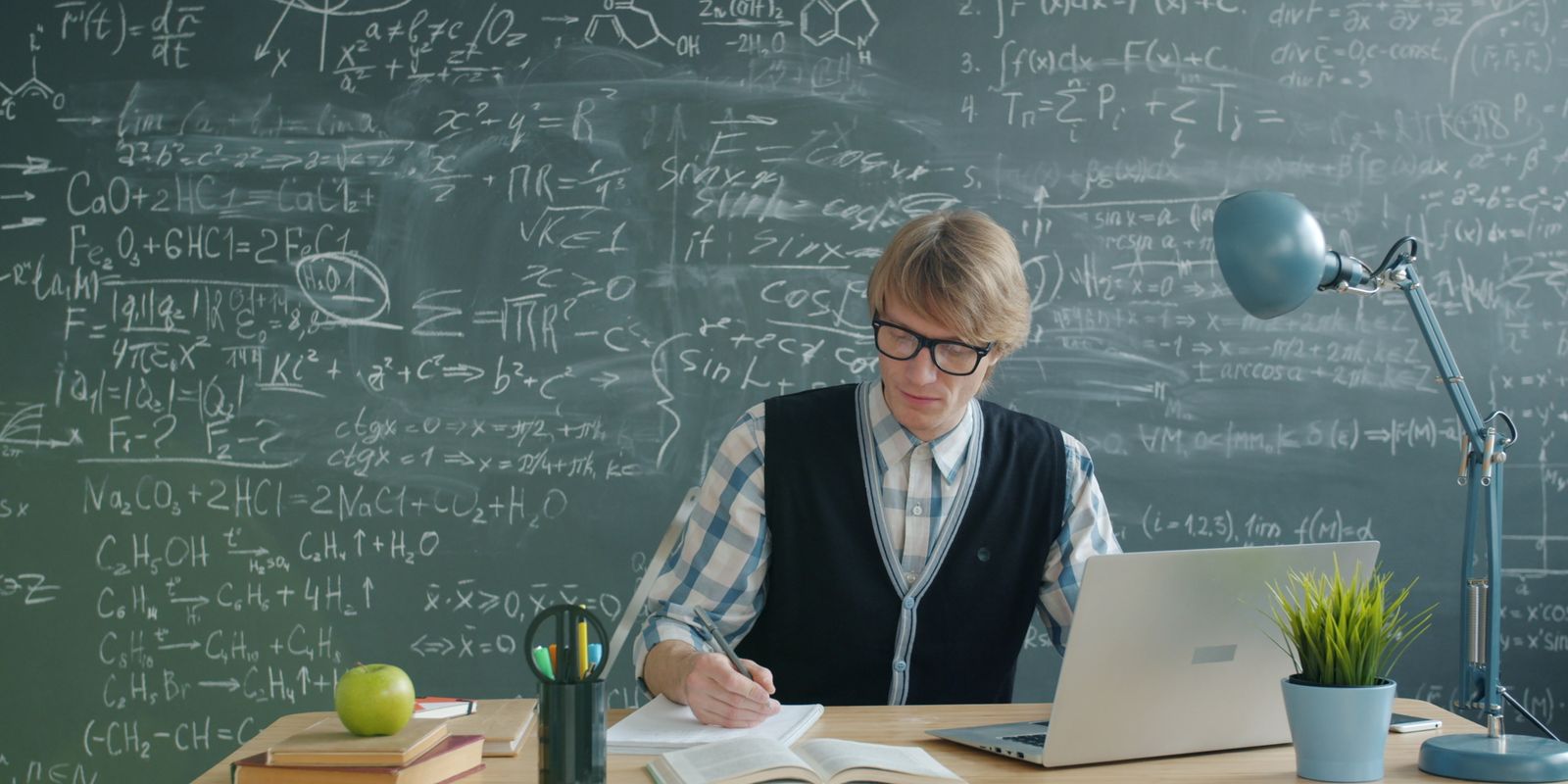 A teacher writing lesson notes at a desk in front of a chalkboard, with a laptop open beside them